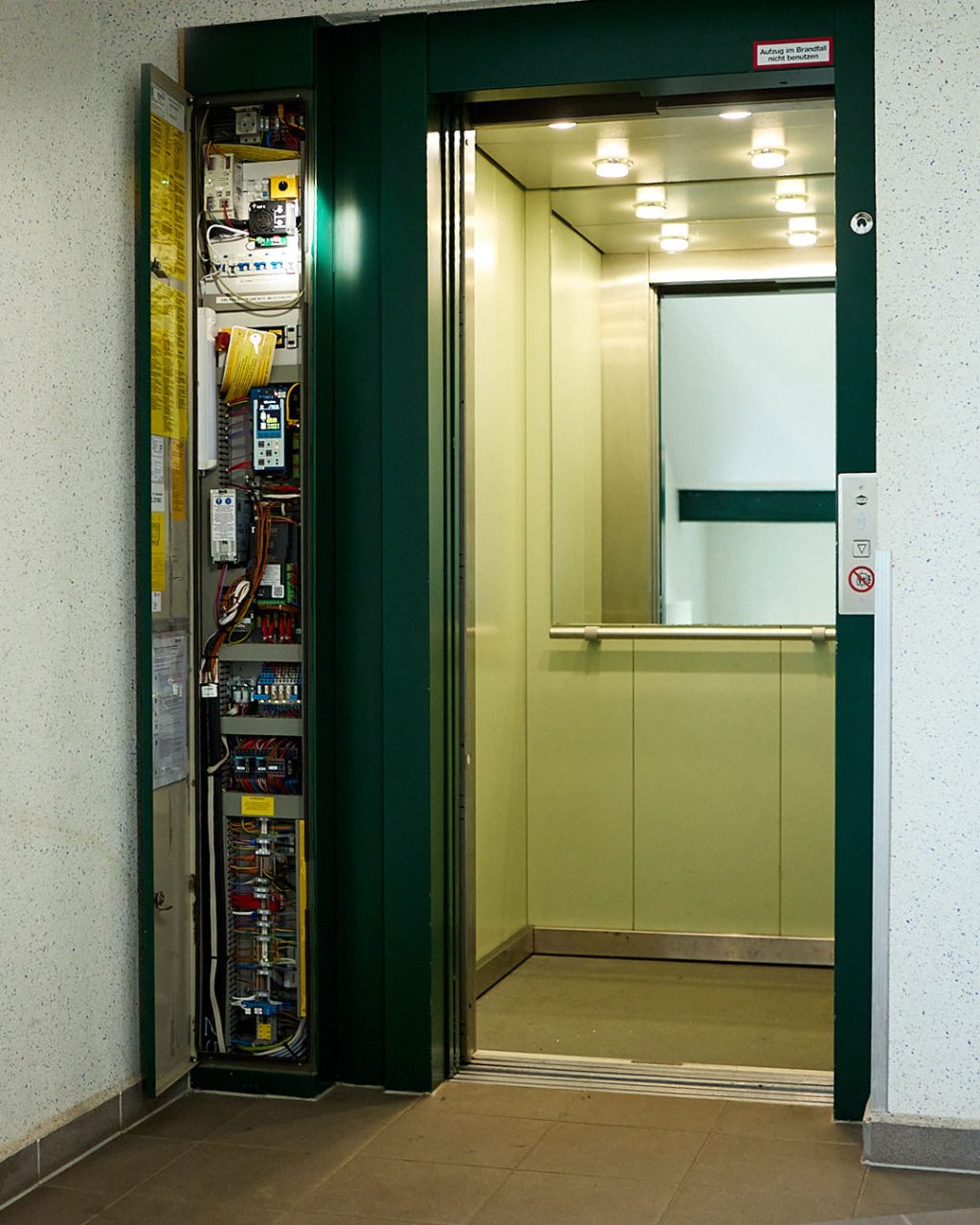 Aufzugsanlage im Wartungsmodus: Geöffnete grüne Tür neben dem freigelegten Schaltschrank mit Steuerung und Verkabelung