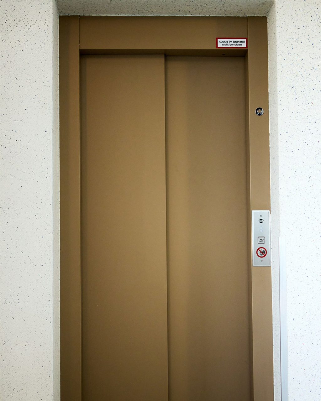 Geschlossene, bronzefarbene Aufzugstür mit Außen-Bedienpanel und Brandschutzhinweis ('Aufzug im Brandfall nicht benutzen')