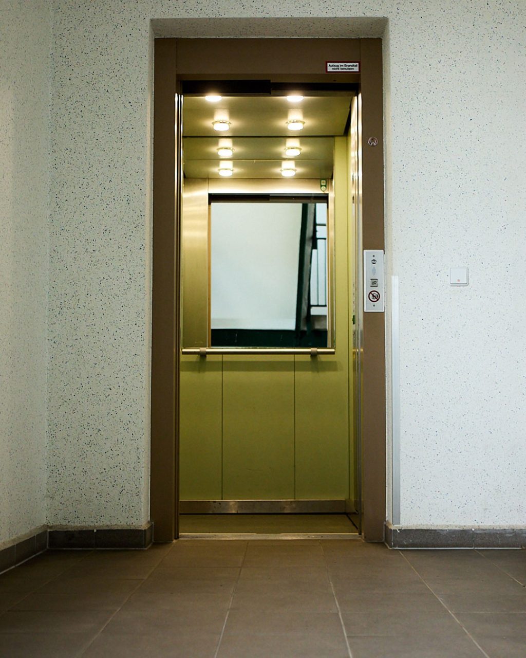 Geöffnete Aufzugtür in beiger Verkleidung, bietet einen klaren Blick in die helle Kabine mit Spiegel und Beleuchtung
