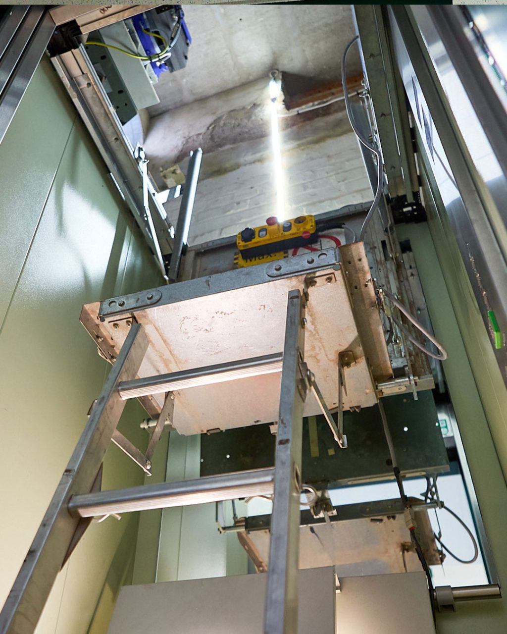 Blick von unten in den Aufzugsschacht auf die Montageplattform über der Kabine, mit Wartungsschalter und Leiter