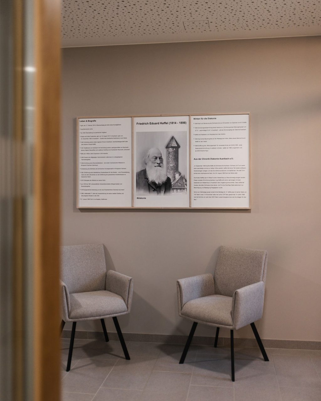 Infotafel an einer hellen Wand im Keffelhaus über das Leben und Wirken von Friedrich Eduard Keffel (1814–1898) mit einer historischen Porträtaufnahme und Textspalten zur Diakonie Auerbach. Davor stehen zwei moderne, hellgraue Polstersessel mit schwarzen Stuhlbeinen auf grauem Fliesenboden.