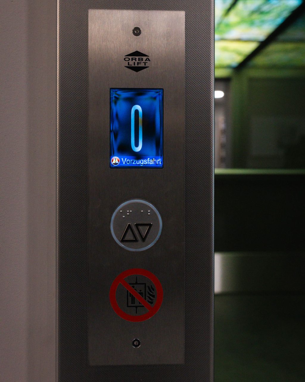 Frontale Nahaufnahme des äußeren Anzeigepanels des Aufzugs. Auf dem bläulich leuchtenden Digitaldisplay steht die Ziffer „0“ sowie der Warnhinweis „Vorzugsfahrt“.