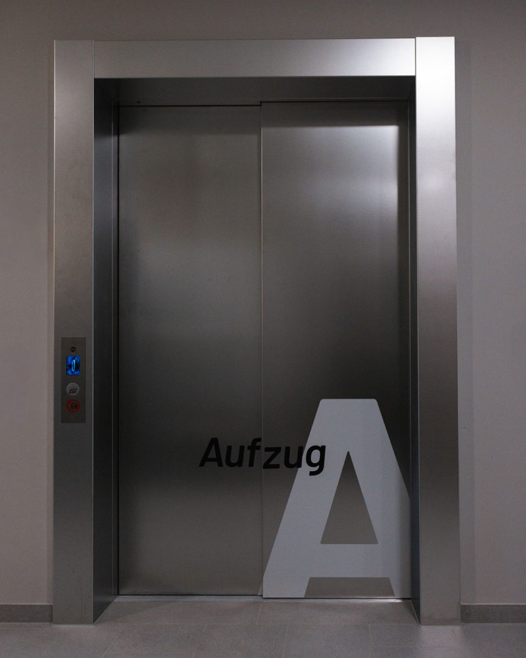 Frontale Ansicht des geschlossenen Aufzugs von außen. Die Schiebetüren aus Edelstahl sind im unteren Bereich mit einer großen, weißen Grafik des Buchstabens „A“ und dem Schriftzug „Aufzug“ versehen. Links befindet sich das schmale Bedientableau.