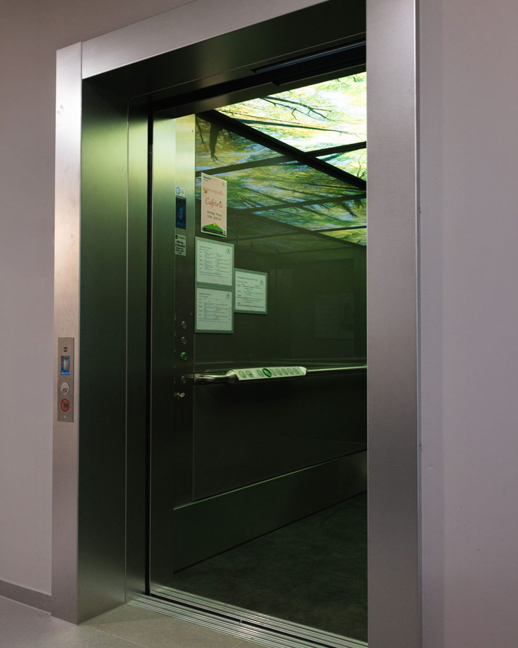 Blick in den geöffneten, modernen Aufzug von Orba Lift. Die Kabine hat Wände aus Edelstahl und eine beleuchtete Decke mit einem großflächigen Fotomotiv grüner Baumkronen im Wald. An den Innenwänden hängen Infoblätter und ein Speiseplan der Cafeteria.