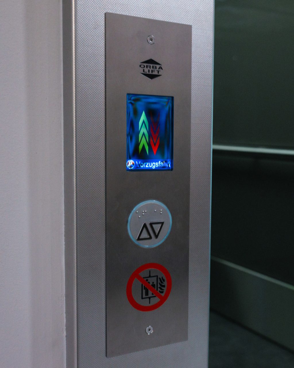 Nahaufnahme des äußeren Bedientableaus aus gebürstetem Edelstahl des Orba Lift Aufzugs. Das digitale Display zeigt farbige Pfeile und den Text „Vorzugsfahrt“. Darunter befinden sich die Ruftaste mit Brailleschrift und ein Verbotszeichen für die Nutzung im Brandfall.