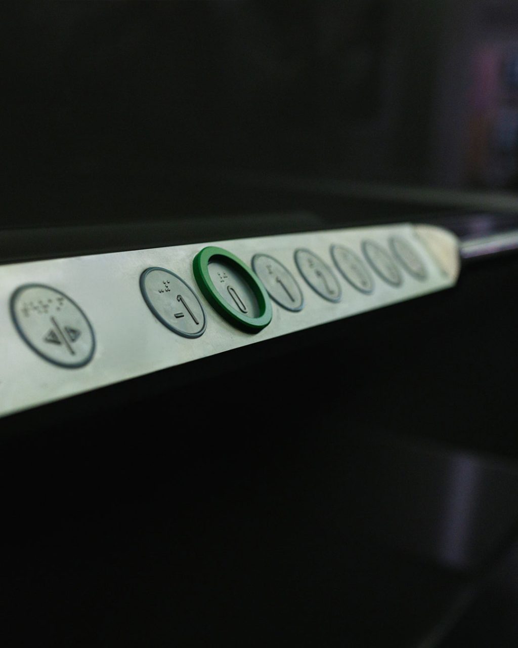Detailaufnahme der horizontalen Tastenleiste im Inneren des Aufzugs. Die Tasten sind mit erhabenen Zahlen und Brailleschrift für blinde Menschen versehen. Die Taste für das Erdgeschoss (0) ist mit einem auffälligen, grünen Ring markiert.