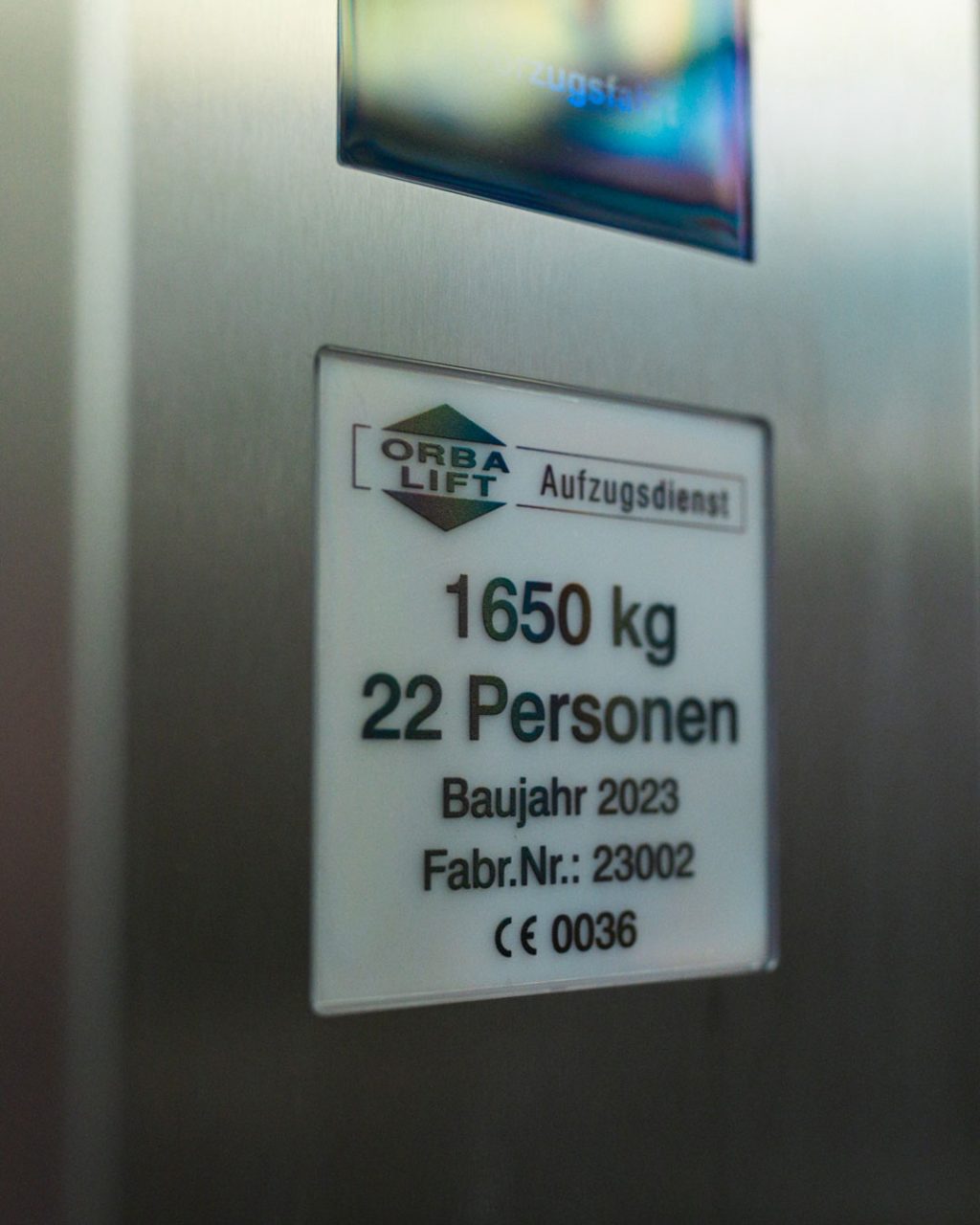 Nahaufnahme des Herstellerschilds von „ORBA LIFT Aufzugsdienst“ an der Aufzugswand. Es listet die technischen Daten auf: Tragkraft 1650 kg, Kapazität für 22 Personen, Baujahr 2023, Fabriknummer 23002 und das CE-Zeichen mit der Nummer 0036.
