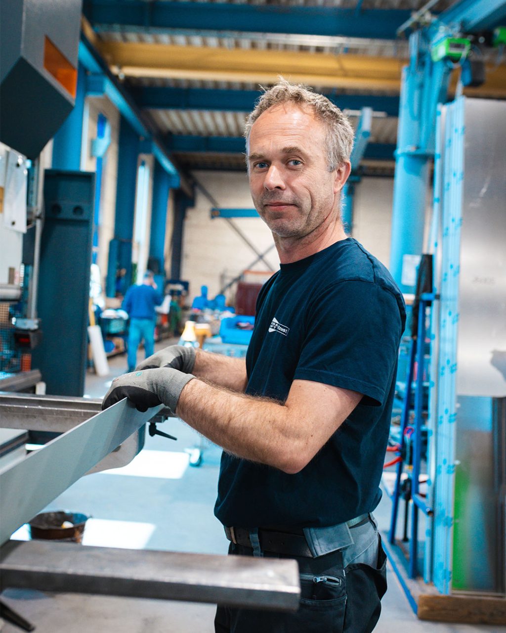 Ein Metallbauer schaut lächelnd in die Kamera, während er ein Blech an einer Maschine bearbeitet.