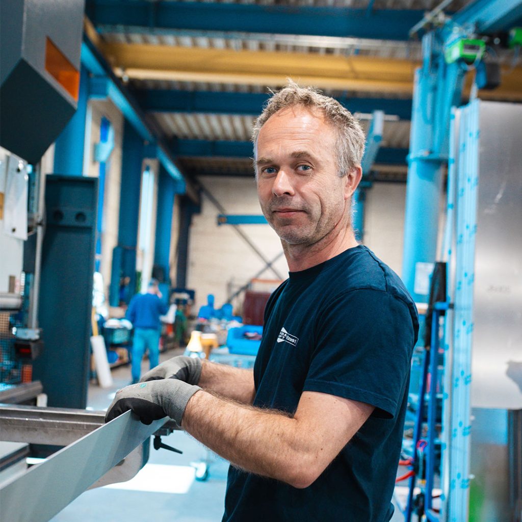 Ein Metallbauer schaut lächelnd in die Kamera, während er ein Blech an einer Maschine bearbeitet.