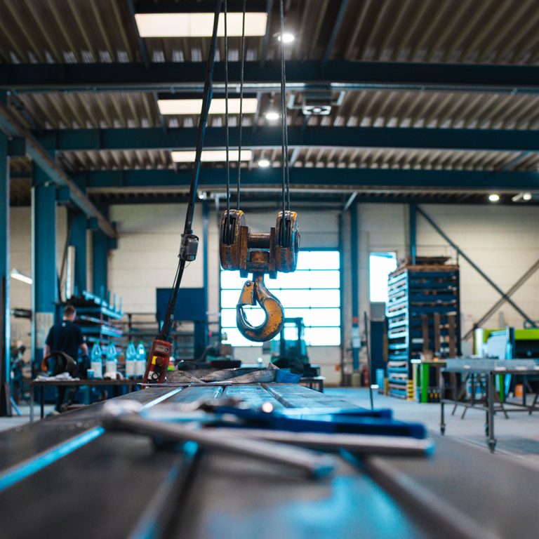 Gelber Industriekranhaken an Stahlseilen, der über einem Metallarbeitstisch in einer Industriehalle von ORBA-Lift hängt.