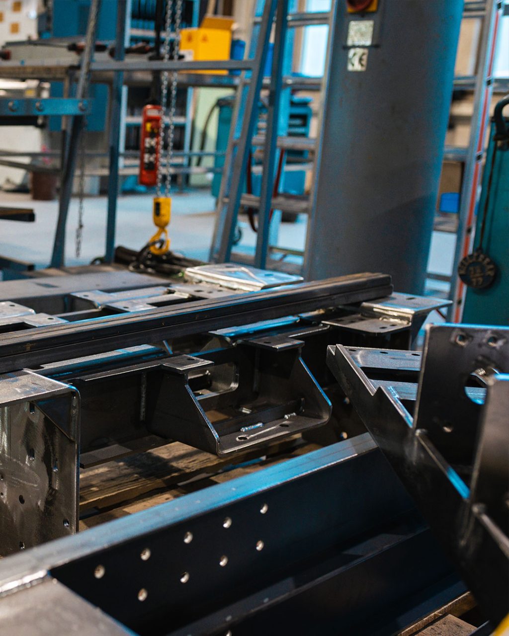 Detailansicht von massiven Stahlkomponenten und Trägern auf einer Holzpalette in einer Produktionshalle, im Hintergrund sind Werkzeuge und Industrieregale zu erkennen.