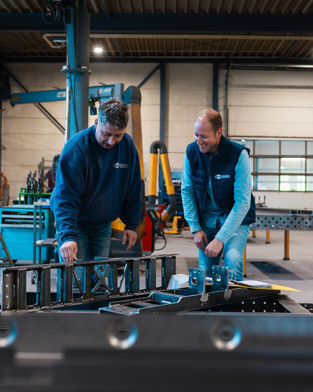 Zwei Mitarbeiter in einer Werkstatt stehen in einer Werkhalle vor einem Arbeitstisch und begutachten und besprechen gemeinsam gefertigte Metall- und Stahlelemente.
