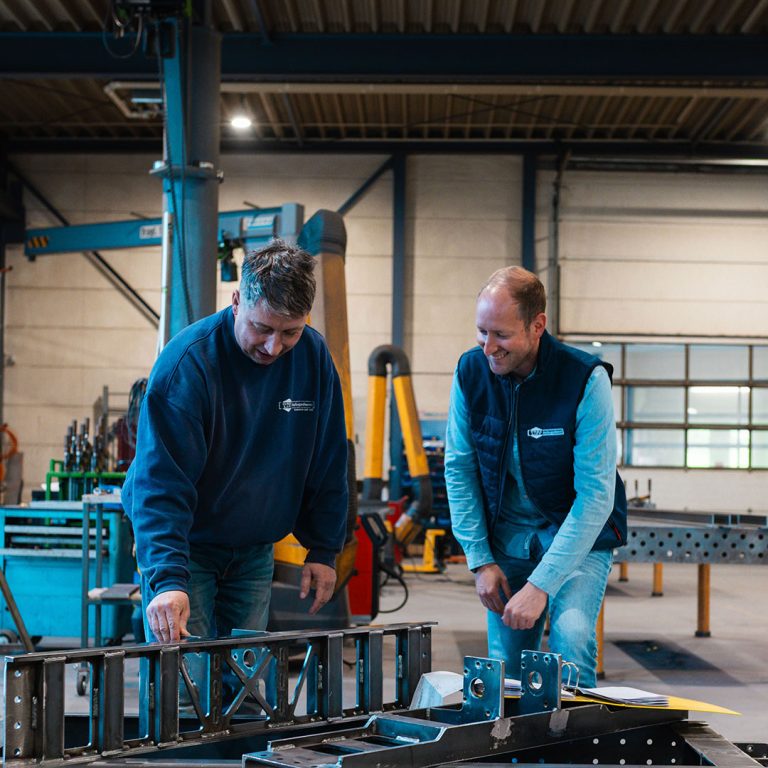 Zwei Mitarbeiter in einer Werkstatt stehen in einer Werkhalle vor einem Arbeitstisch und begutachten und besprechen gemeinsam gefertigte Metall- und Stahlelemente.