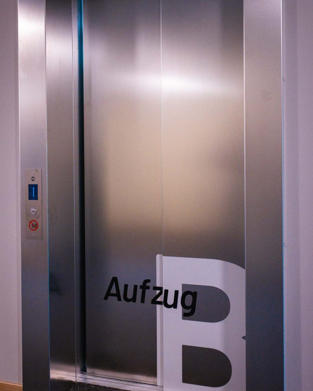 Leicht seitliche Nahaufnahme eines Aufzugs mit geschlossenen Metalltüren. Im unteren Bereich steht der Schriftzug „Aufzug B“. Links im silbernen Rahmen befindet sich das schmale Tableau mit blauer Etagenanzeige und Tastern.