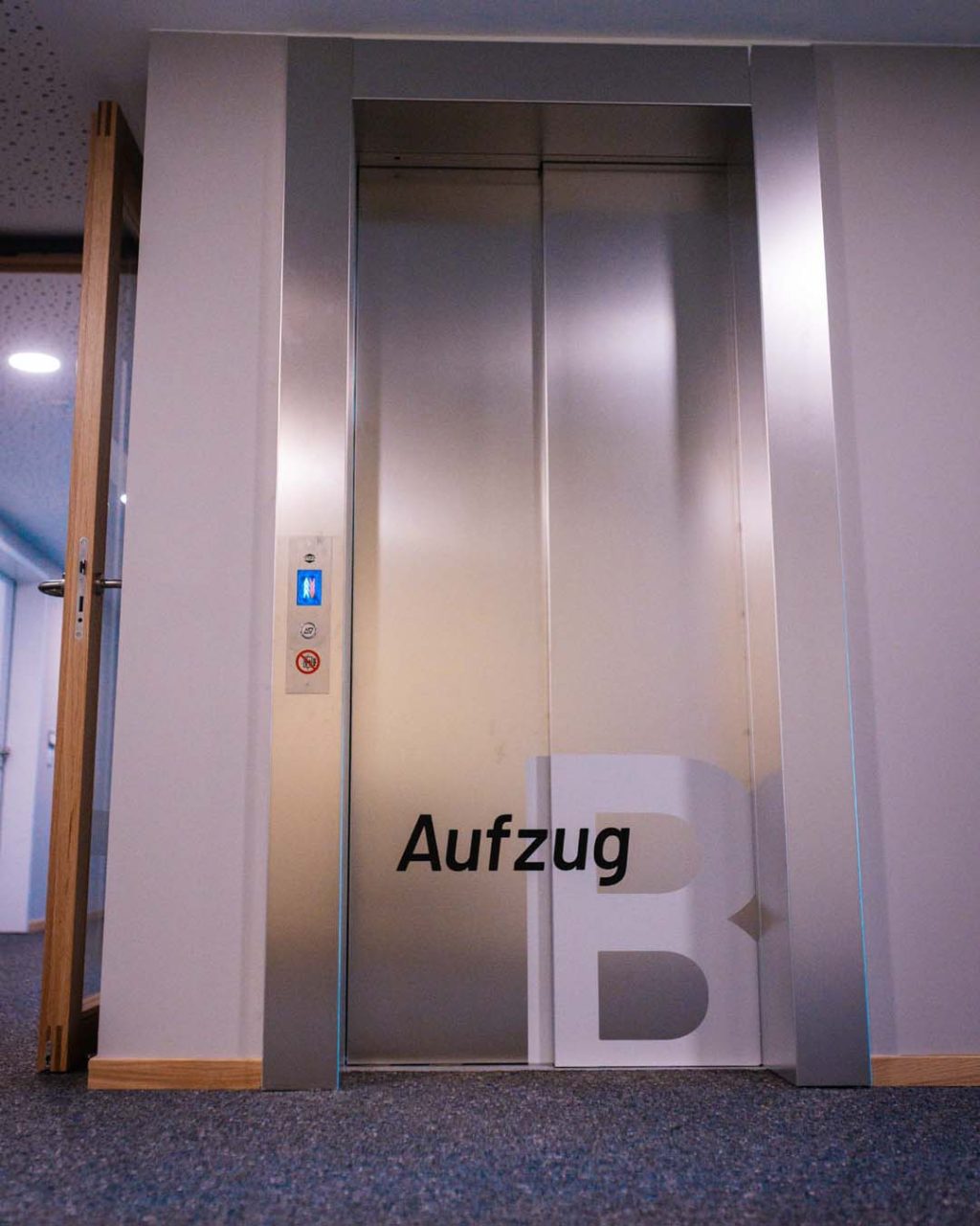 Frontalansicht eines modernen Aufzugs aus gebürstetem Edelstahl mit geschlossenen Türen. Auf den Türen befindet sich unten die dunkle Aufschrift „Aufzug B“. Links am Rahmen ist das digitale Bedienfeld zu sehen.