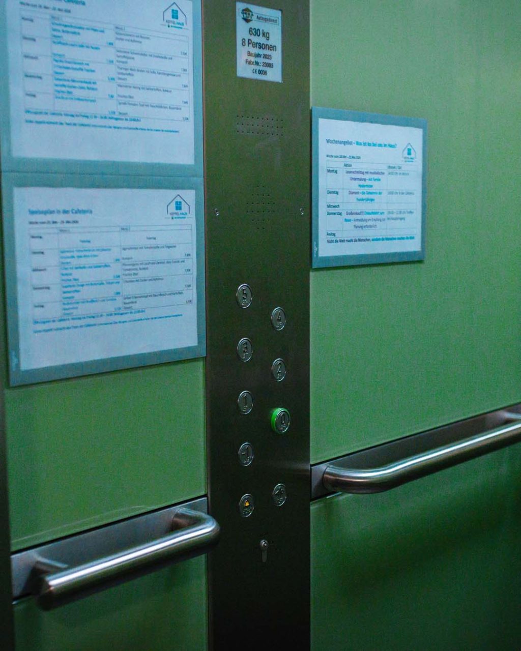 Blick in den Aufzug auf das vertikale Edelstahl-Bedienfeld mit nummerierten Etagentasten. An der grünen Innenwand sind links Speisepläne und rechts ein Wochenangebot in Klarsichthüllen angebracht. Ein metallener Haltegriff verläuft waagerecht durch das Bild.