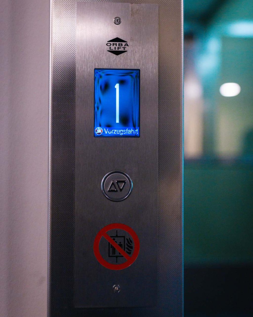 Detailansicht des metallenen Bedientableaus neben der Aufzugstür. Das blaue Display zeigt die Ziffer „1“ und den Schriftzug „Vorzugsfahrt“. Darunter befinden sich ein runder Taster sowie ein Rot-Weißes Piktogramm, das die Nutzung im Brandfall verbietet.