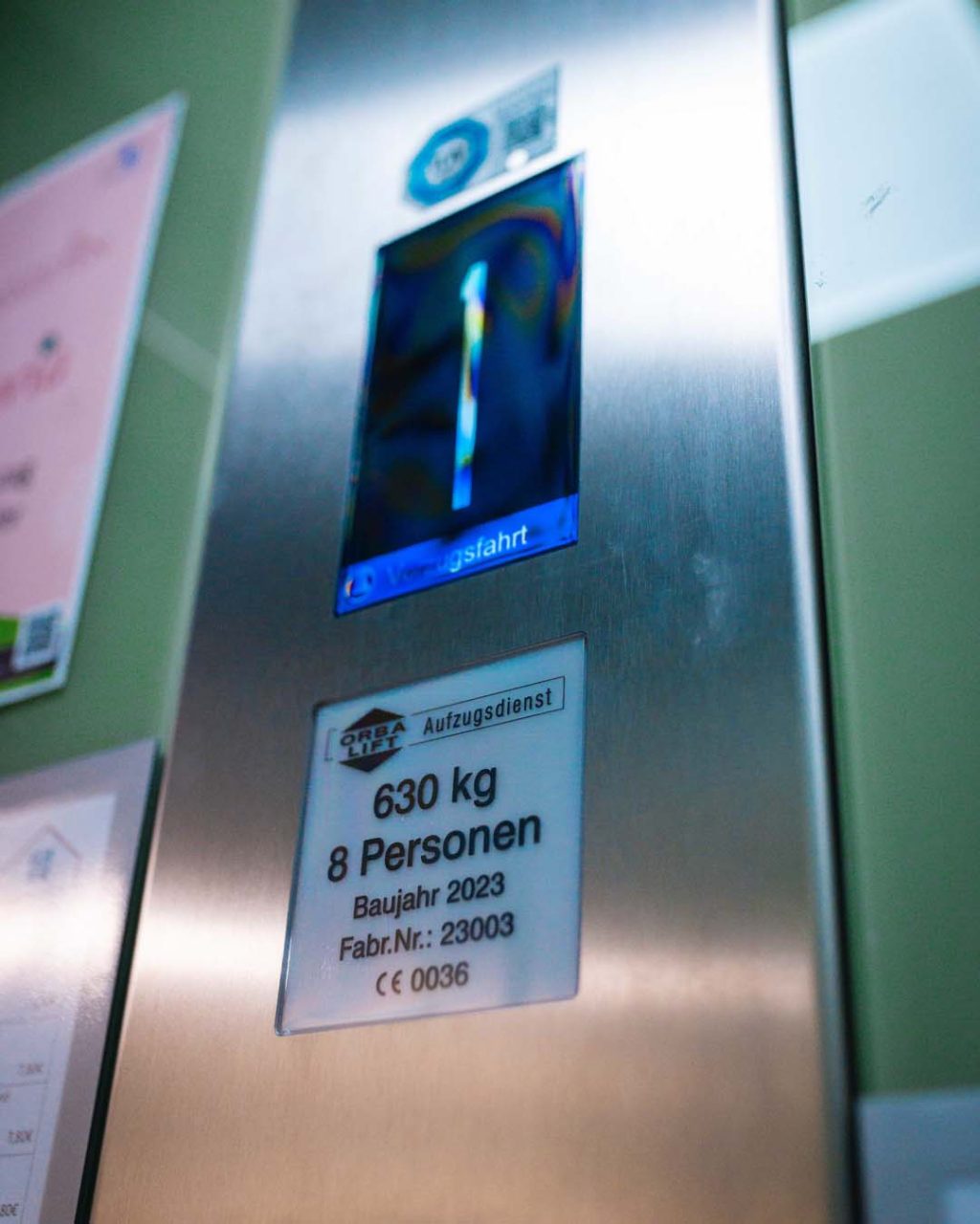 Nahaufnahme des internen Aufzugspaneels. Auf einem Schild steht „ORBA LIFT Aufzugsdienst“, „630 kg / 8 Personen“ sowie das Baujahr 2023. Darüber zeigt die blaue Digitalanzeige die Stockwerkszahl 1.