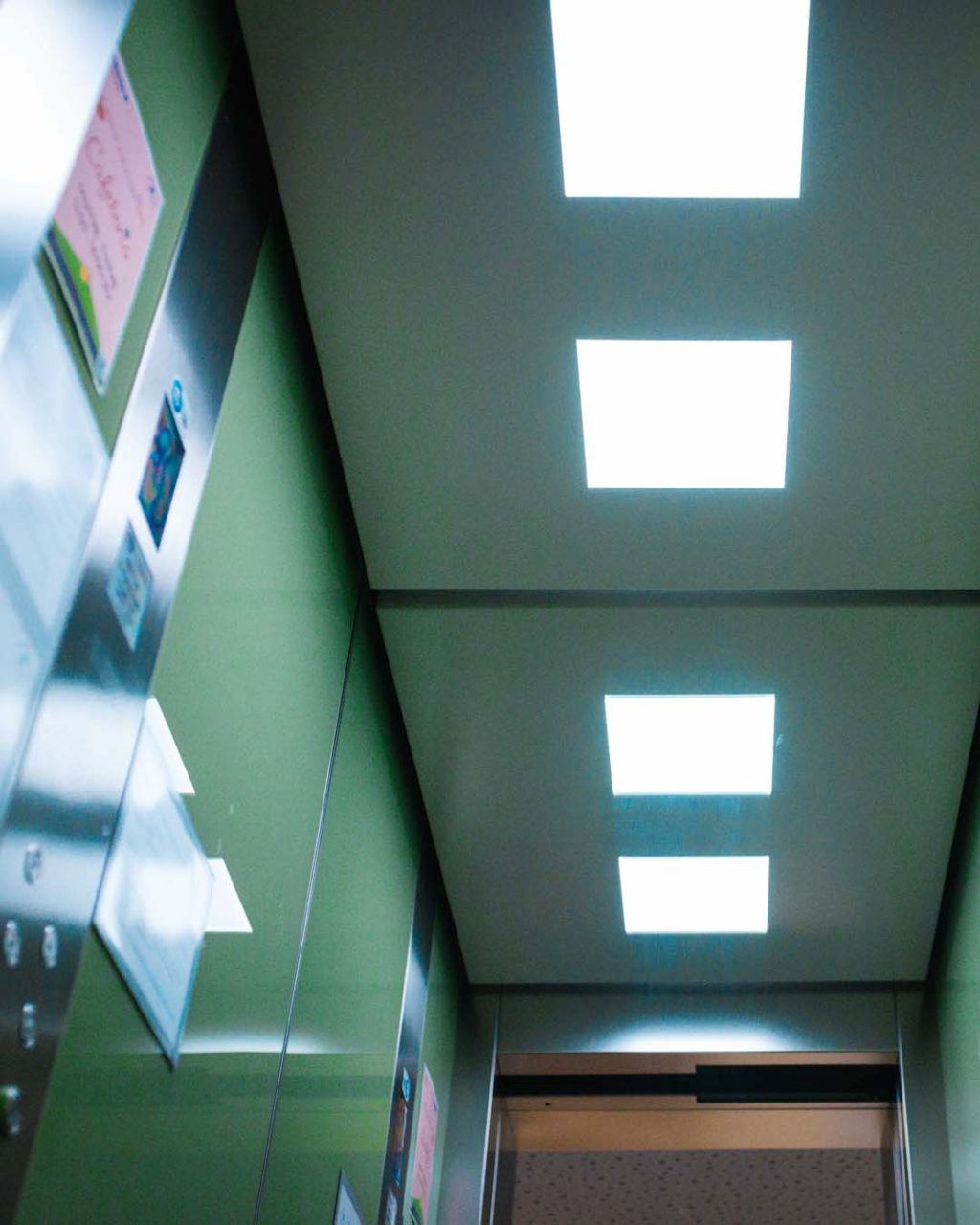 Froschperspektive im Inneren des Aufzugs mit Blick auf die Deckenbeleuchtung, bestehend aus vier leuchtenden, quadratischen LED-Panels. Links ist die grüne Wand mit Aushängen zu erkennen.