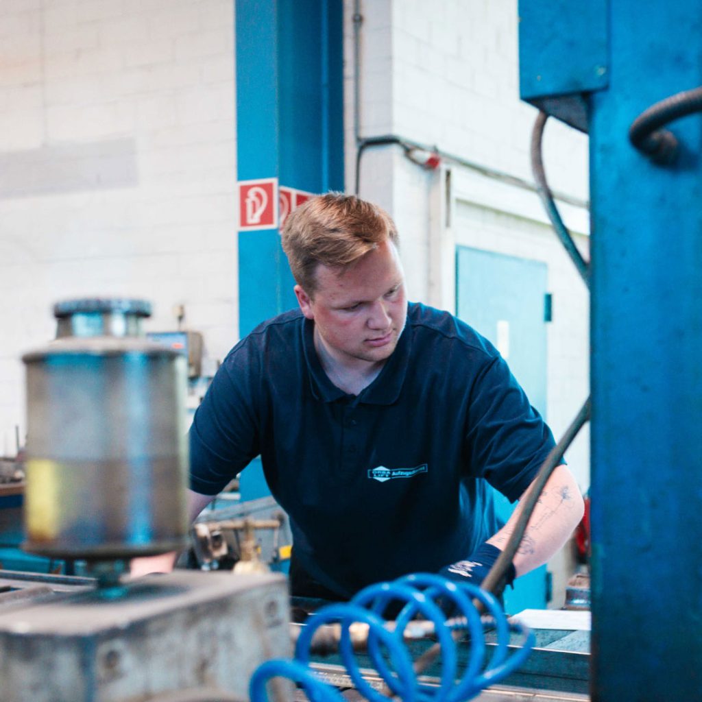 Ein Handwerker in dunkelblauem Arbeitshemd betrachtet aufmerksam eine Arbeitsfläche in einer Werkstatt.