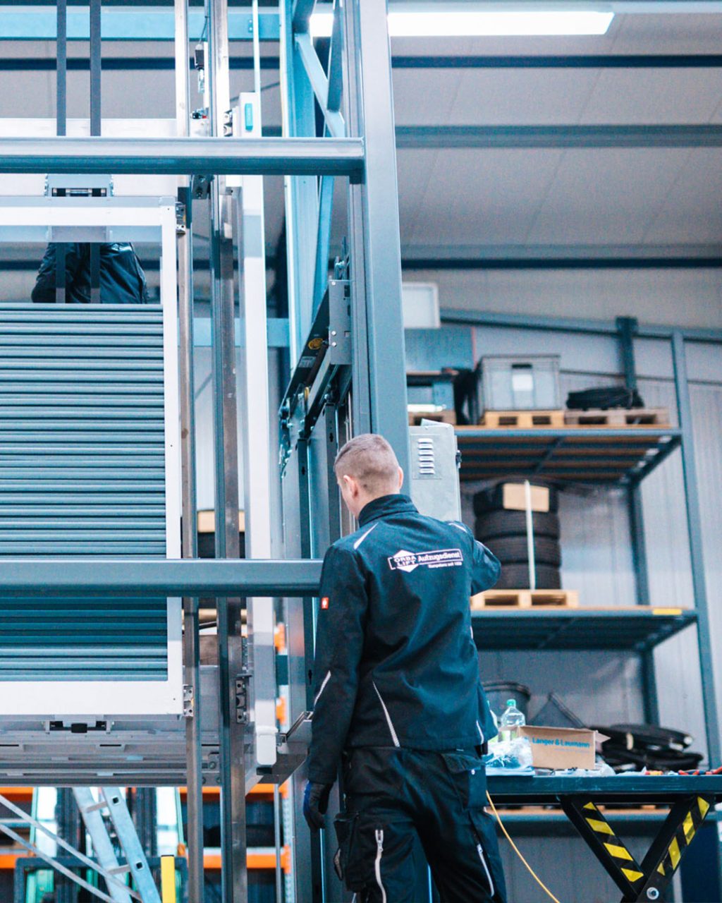 Techniker von ORBA-Lift steht von hinten gesehen in einer Werkstatt vor einer Aufzugskonstruktion und führt Arbeiten durch.